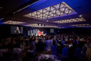 Large conference hall with geometric ceiling lights and crowd seated at round tables during evening event.