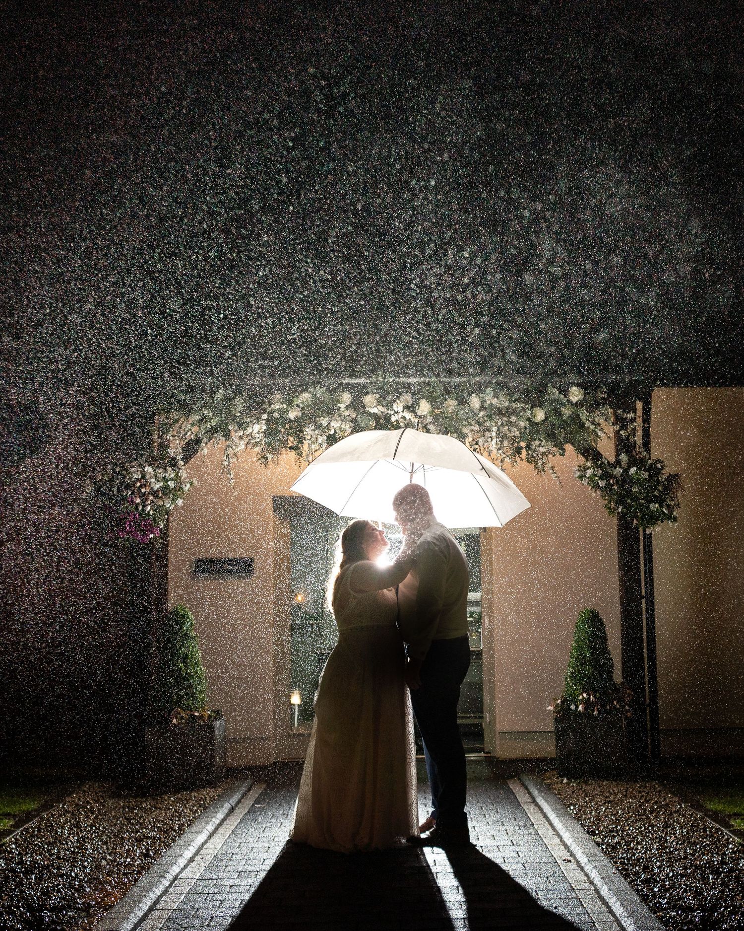 Rainy night shot of a bride and groom under an umbrella, backlit by flash.