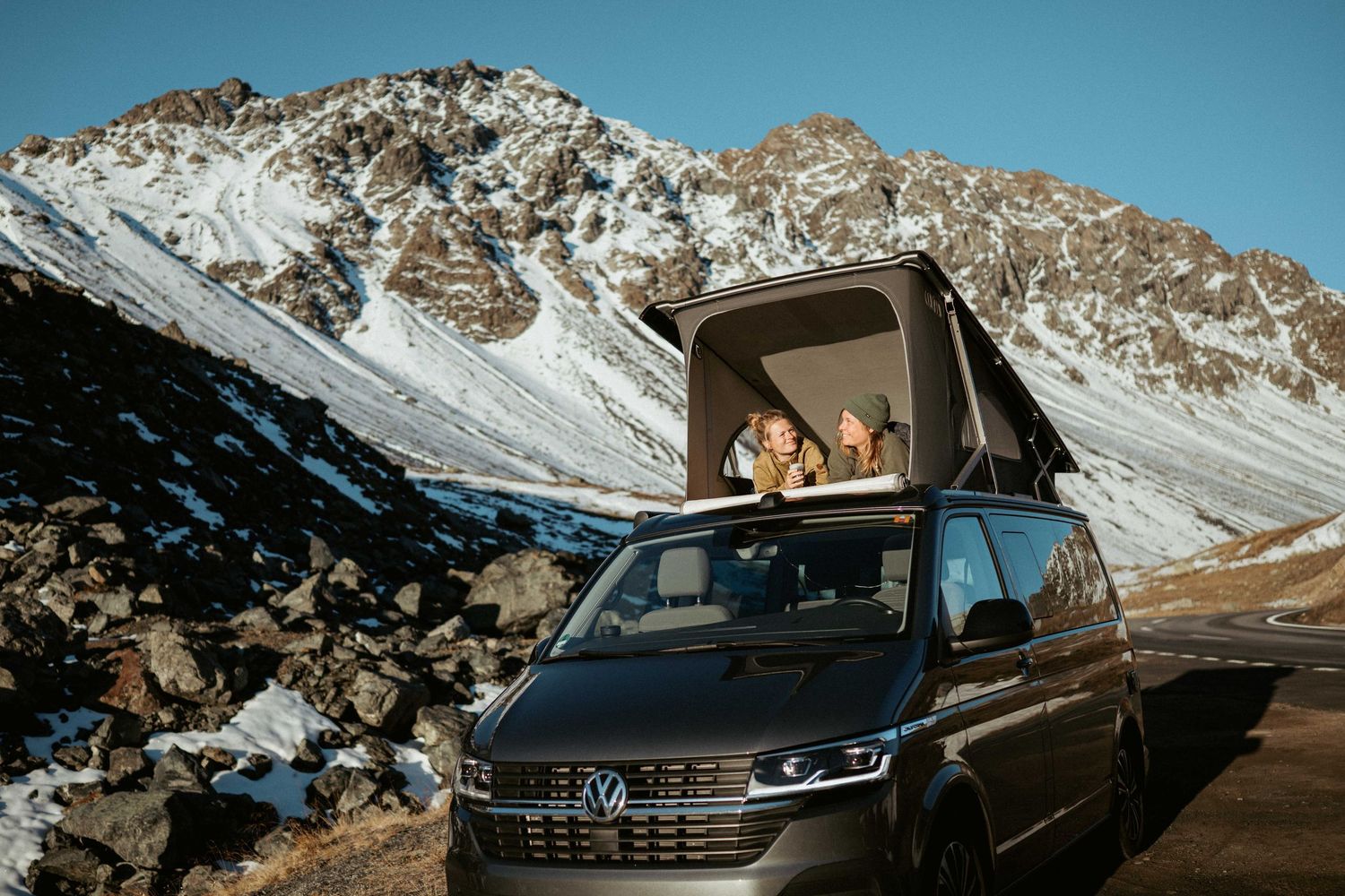 A black Volkswagen SUV drives through a snowy mountain pass with dramatic rocky peaks in the background.