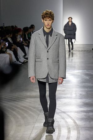 Model walks runway wearing grey blazer and dark pants at fashion show against white backdrop.