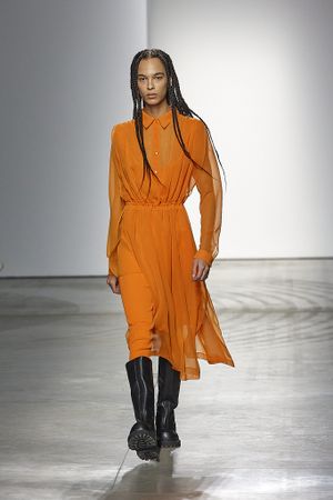 Model showcases flowing orange dress with black boots on fashion runway.