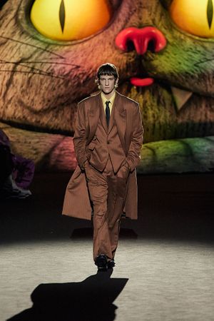Model presents brown suit against dramatic backdrop with large glowing eyes.