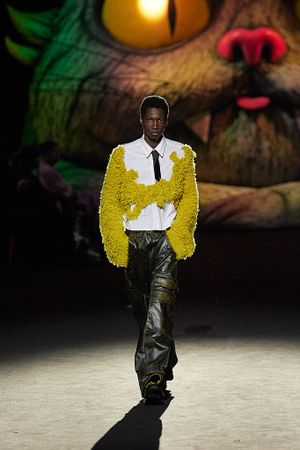 Yellow textured jacket paired with dark pants on catwalk with artistic backdrop.