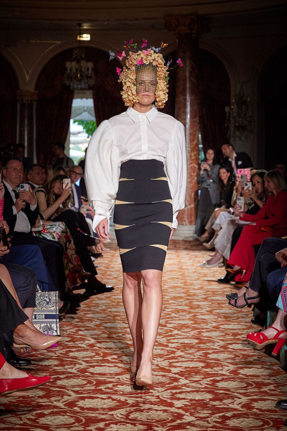 Fashion model walks runway wearing white blouse, striped pencil skirt and floral headpiece at luxury show.