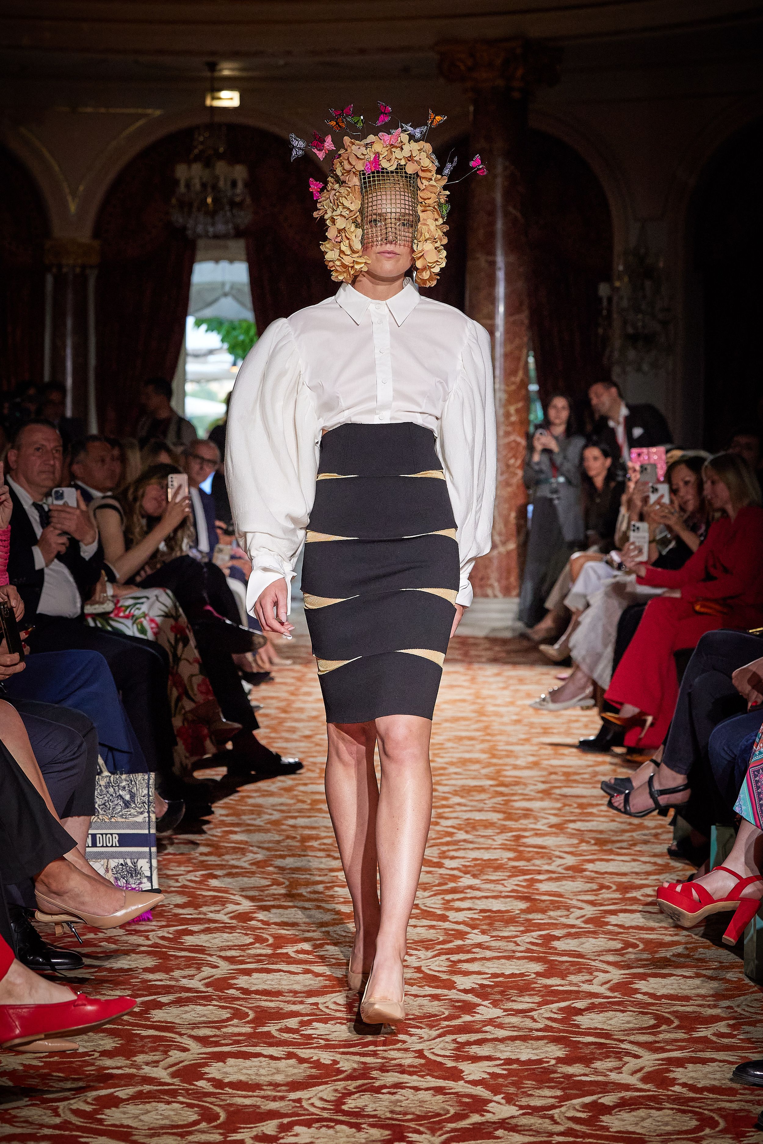 Fashion model walks runway wearing white blouse, striped pencil skirt and floral headpiece at luxury show.