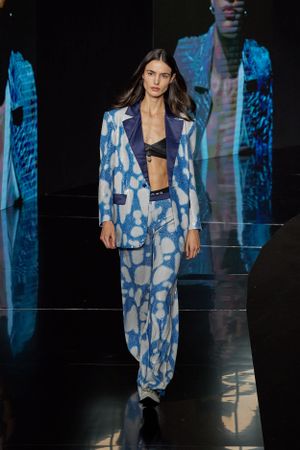 Runway model wearing blue and white patterned pantsuit with crop top on dark catwalk.