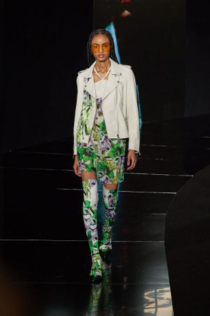 Model presents white leather jacket with floral print pants in high fashion runway show.