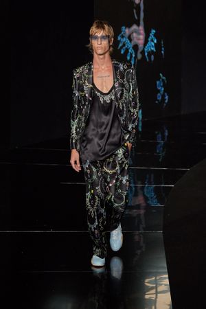 Model wears black silk top with embellished dark patterned suit on runway.