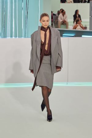 Model wearing oversized pinstriped gray suit with sheer top on modern runway.