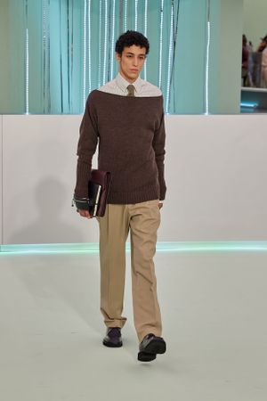 Student-inspired look featuring brown sweater over white collar shirt with khaki pants on runway.