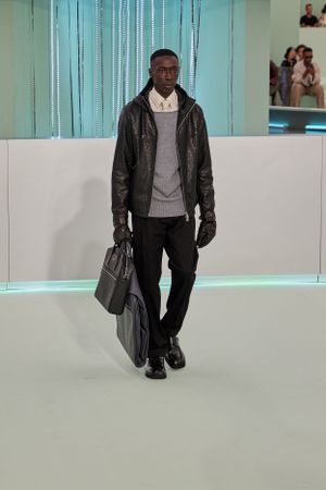 Fashion model wearing dark leather jacket, grey sweater and carrying leather bags on minimalist runway.