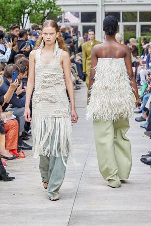 Models showcase fringe-detailed white dresses and wide-leg pants on outdoor runway.
