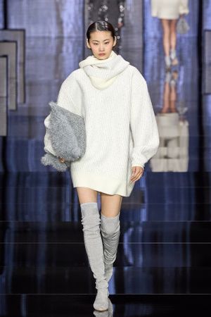 Oversized white knit sweater dress styled with gray thigh-high boots on reflective runway.