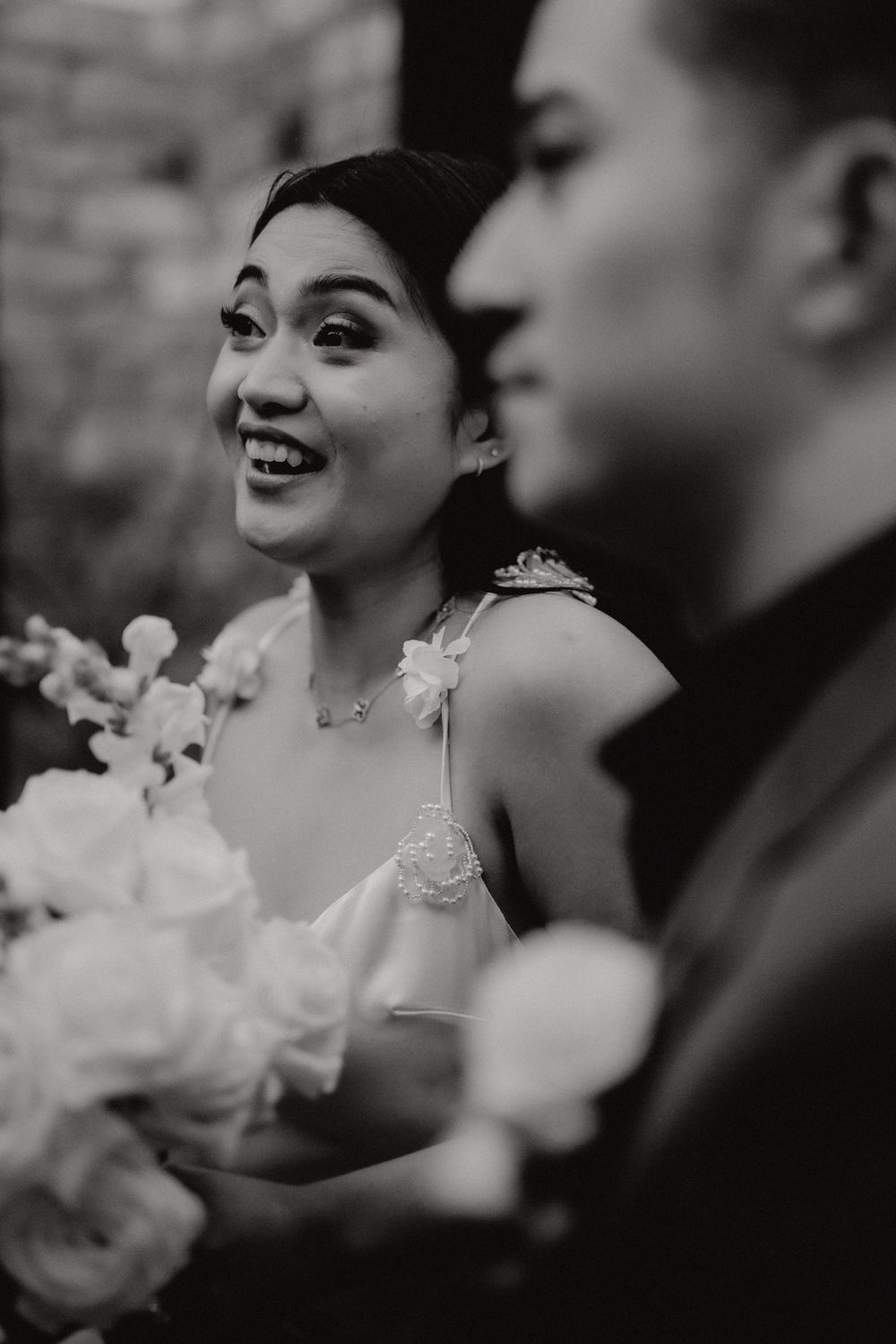 Black and white wedding photo showing a joyful bride in a sleeveless dress holding a bouquet of white flowers.