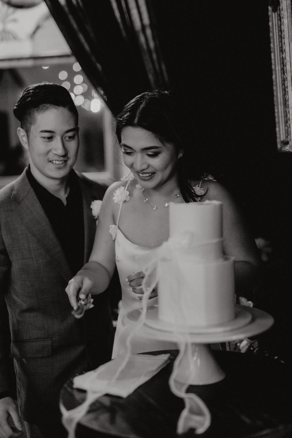 A couple in formal attire smiles while cutting their elegant white wedding cake in a dimly lit setting.