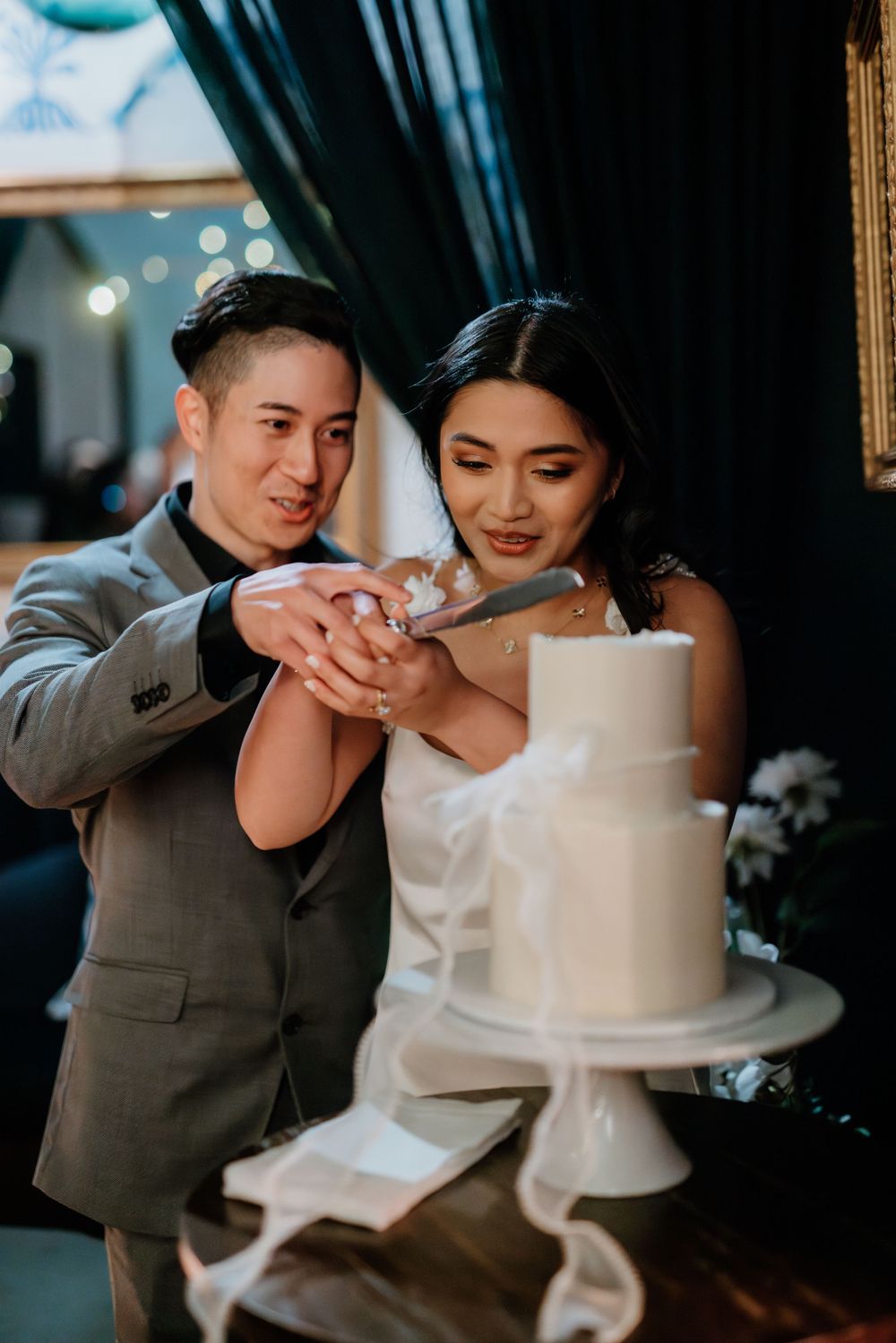 A newlywed couple cuts into their elegant white tiered wedding cake together at their reception celebration.