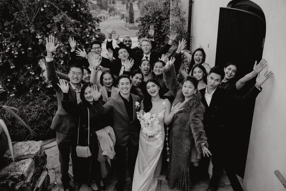 A large wedding party poses joyfully together for a black and white group photo.