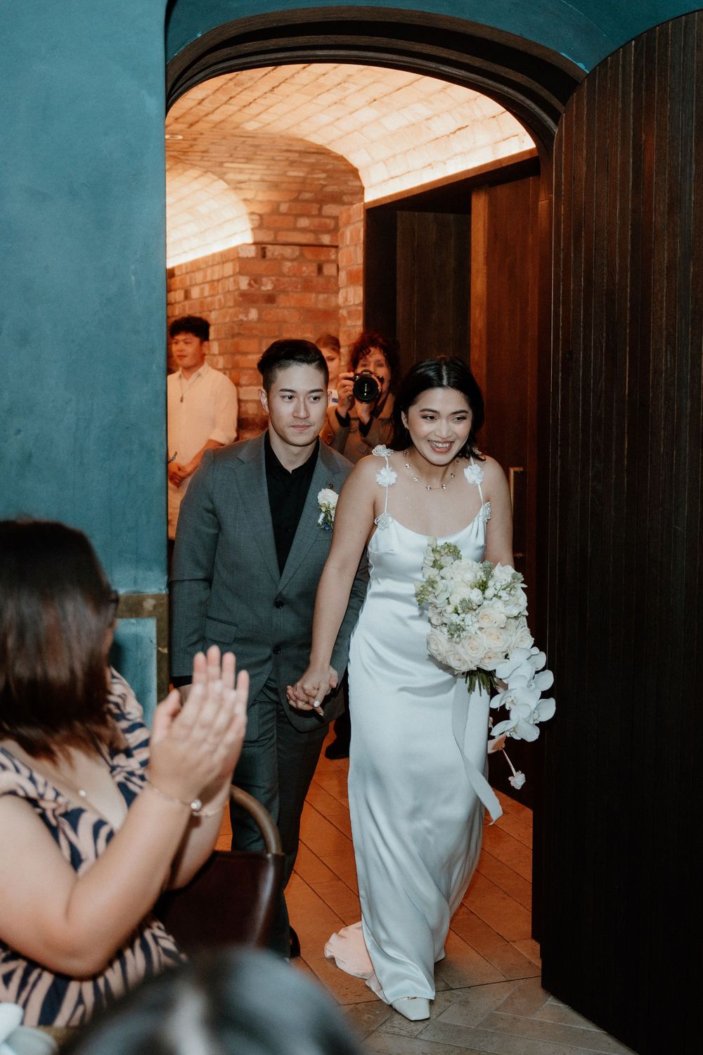 A couple in wedding attire walks through an arched doorway while guests applaud.