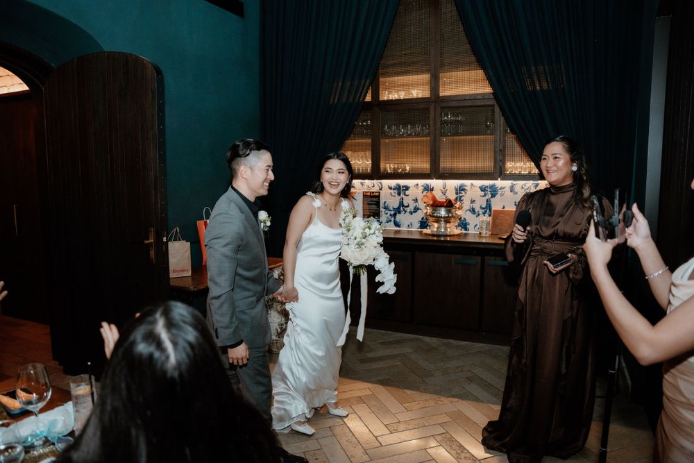 Newlyweds and guests celebrate on a wooden dance floor at an indoor wedding reception with teal curtains in the background.