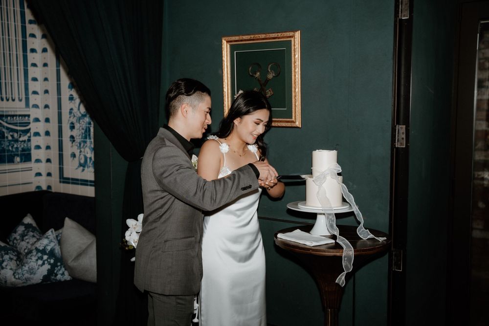 A couple cuts into their elegant white wedding cake together in a room with dark green walls and framed artwork.