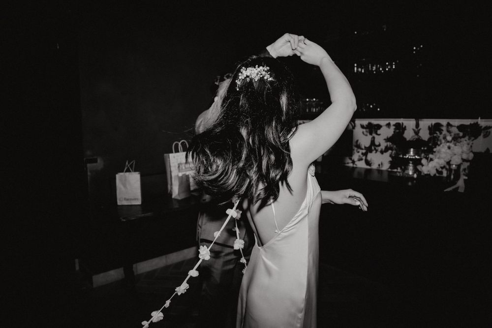 A person in a white dress dances with their arms raised while wearing flowers in their hair at a party.