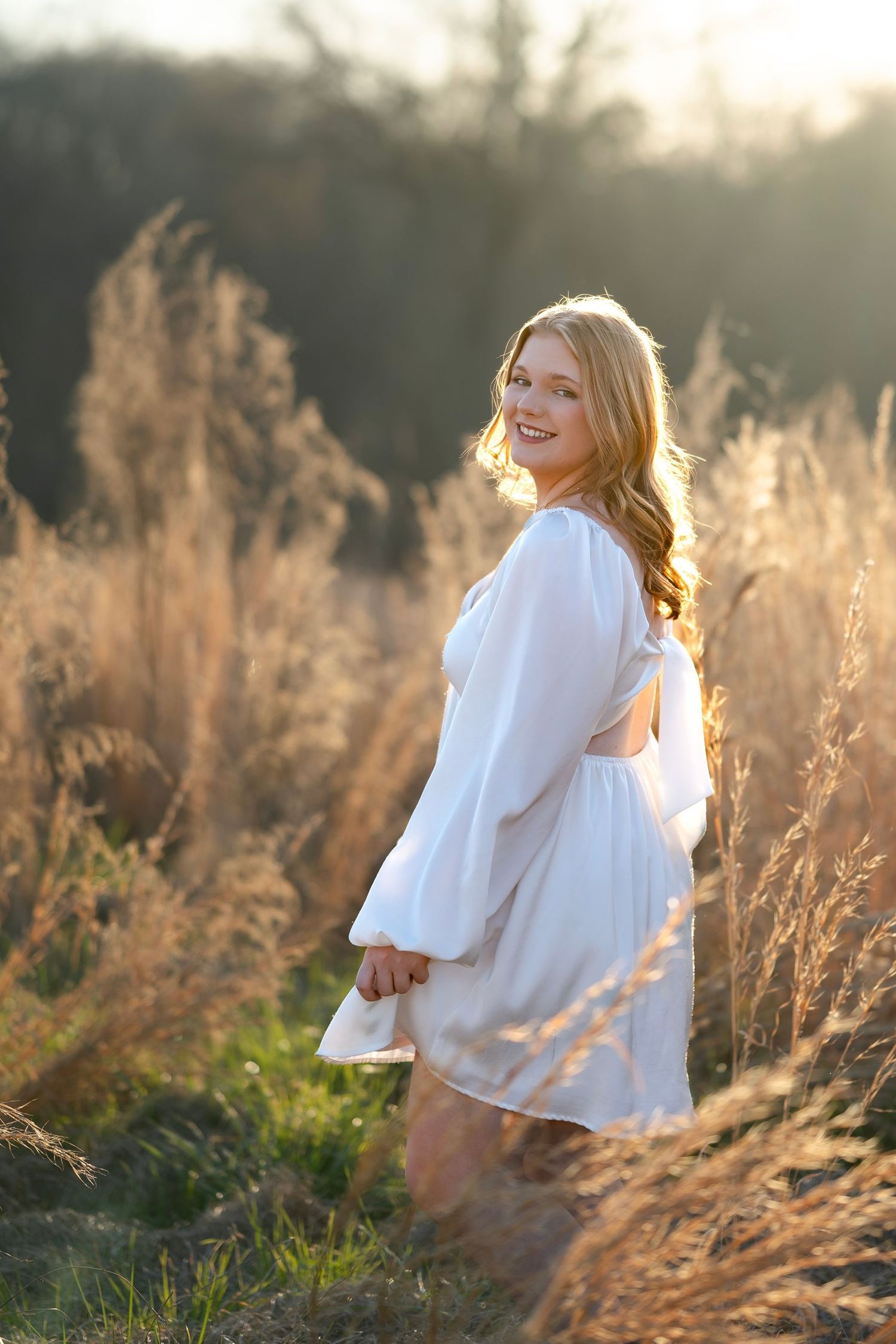 Woman in white dress poses in golden sunlit field of tall grass.