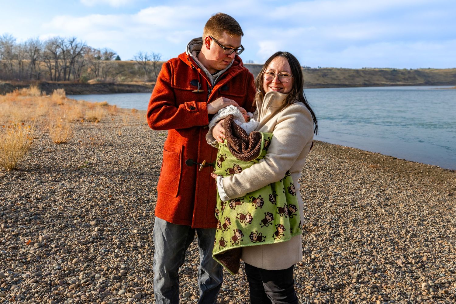 a couple stand on a rocky beach by a river holding their baby.