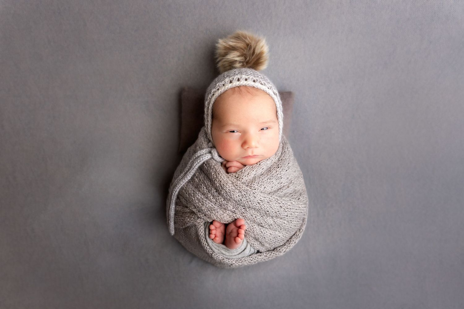 newborn photography session Sammamis in a cozy gray swaddle and knit bonnet lies peacefully on a gray backdrop during a photography session.