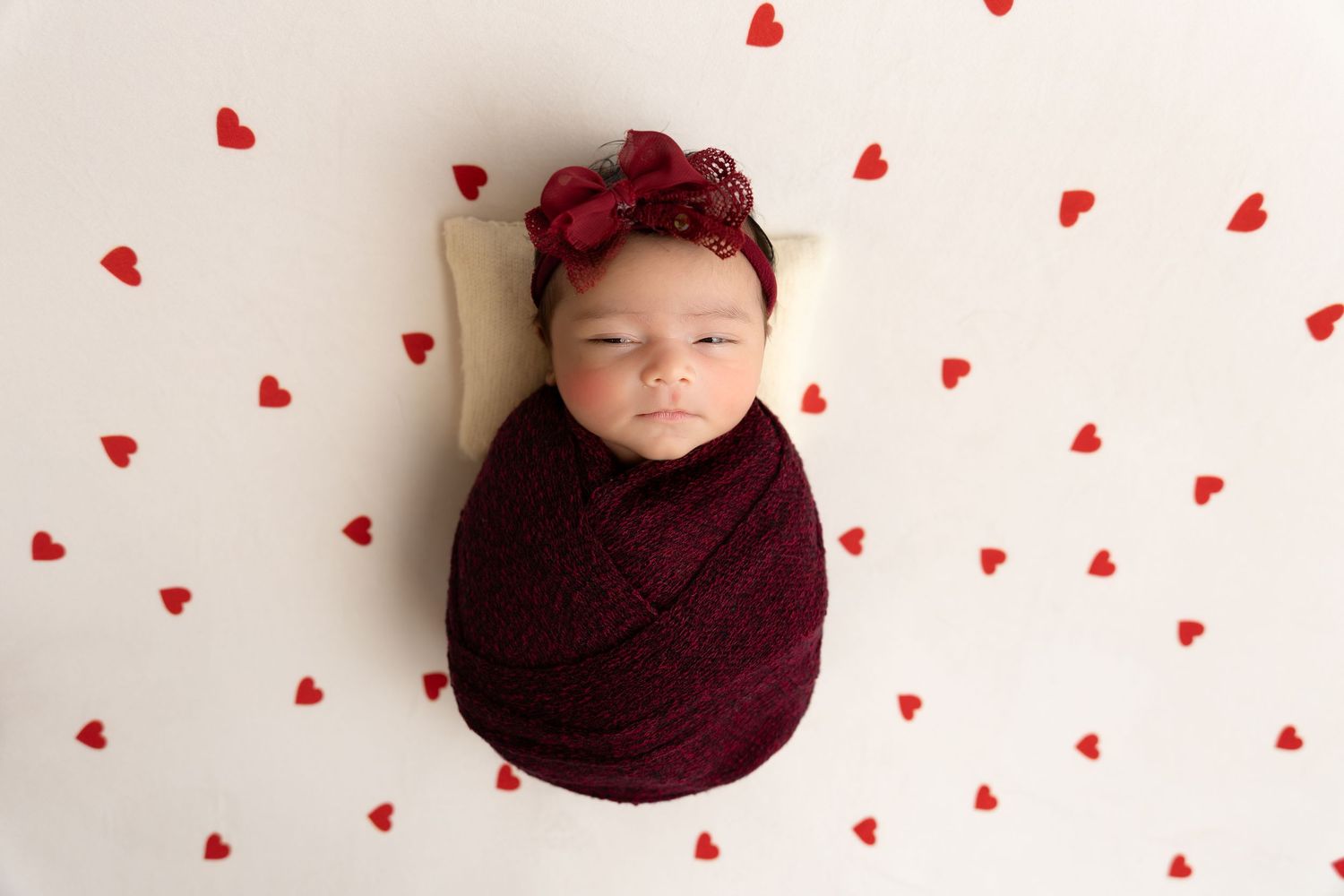 newborn photography session Sammamish WA wrapped in burgundy fabric lies on white background with scattered red paper hearts.