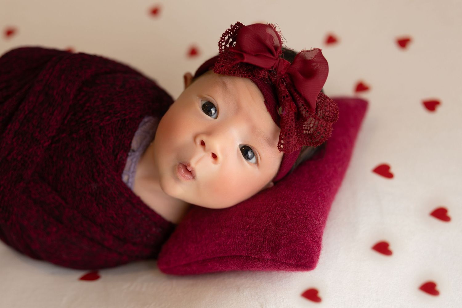 newborn photography session Sammamish WA Baby wearing burgundy bow headband lays on fluffy red blanket surrounded by heart decorations.