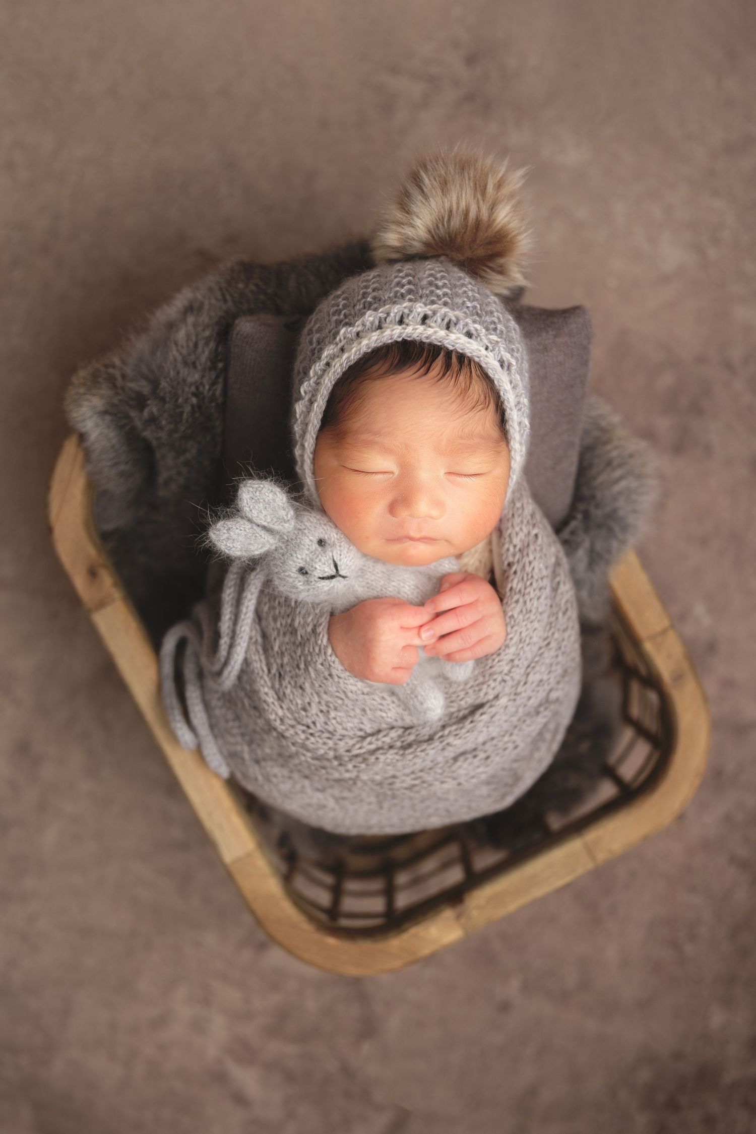 newborn photography session Sammamish WA in cozy gray knit swaddle and hat with pom pom, nestled in wooden basket with fur lining.