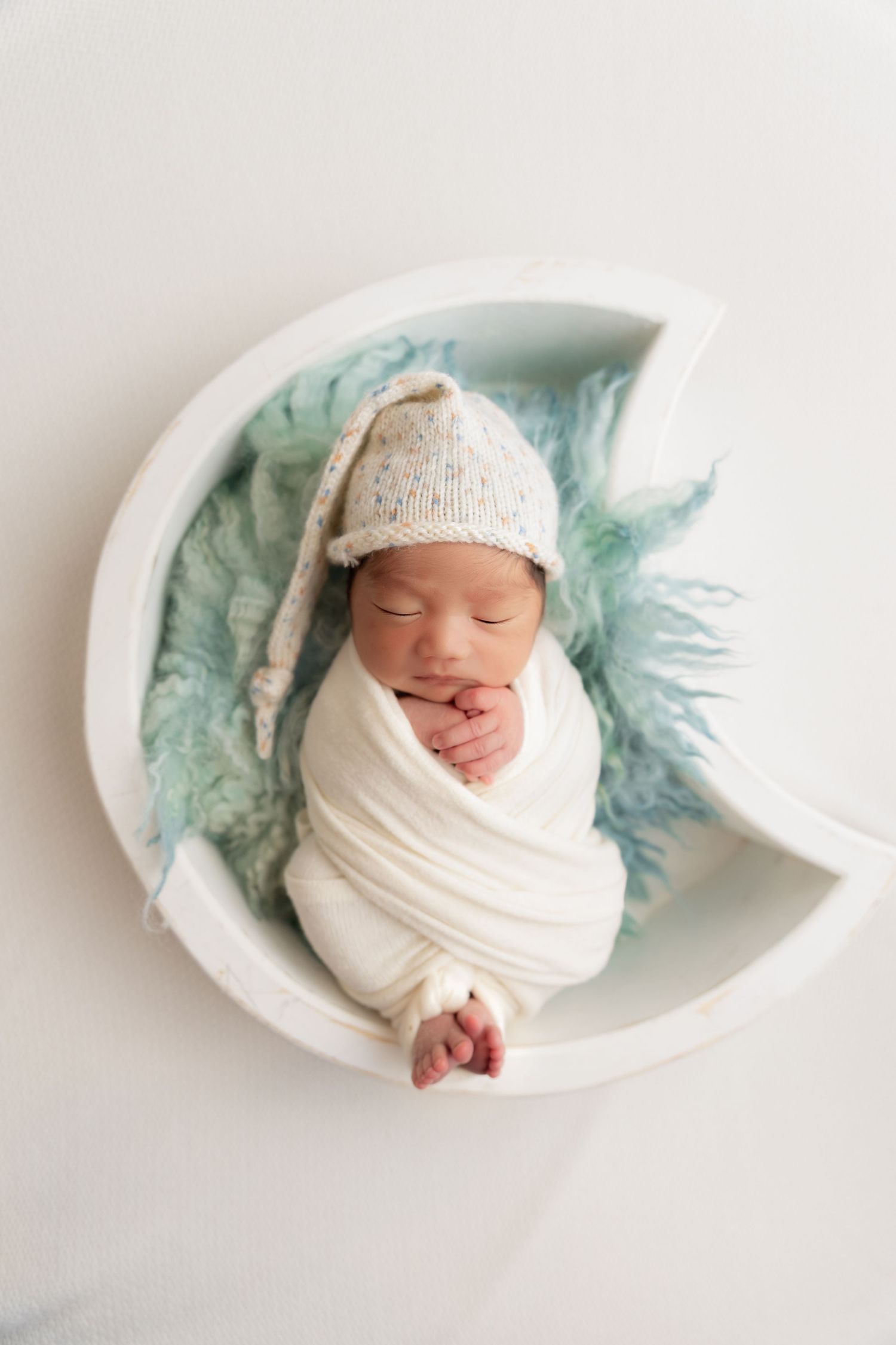 newborn photography session Sammamish WA in white cloth wearing knit hat rests in crescent moon prop with teal fabric backdrop.