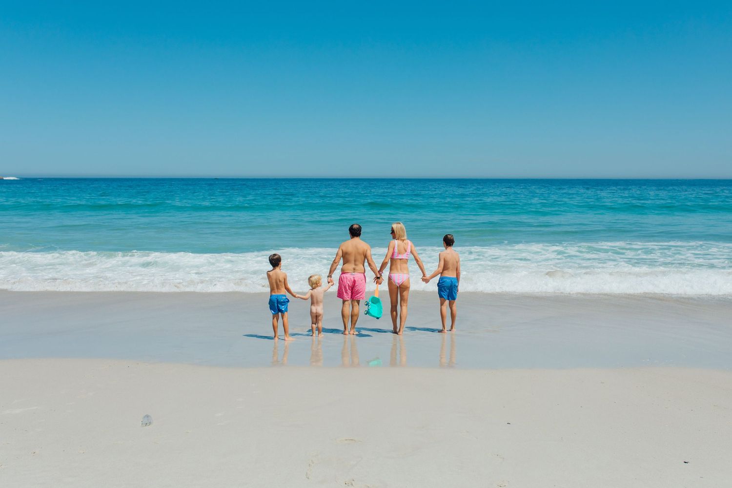 Family of five holding hands on a sunny beach, facing the ocean waves on a clear blue sky day.