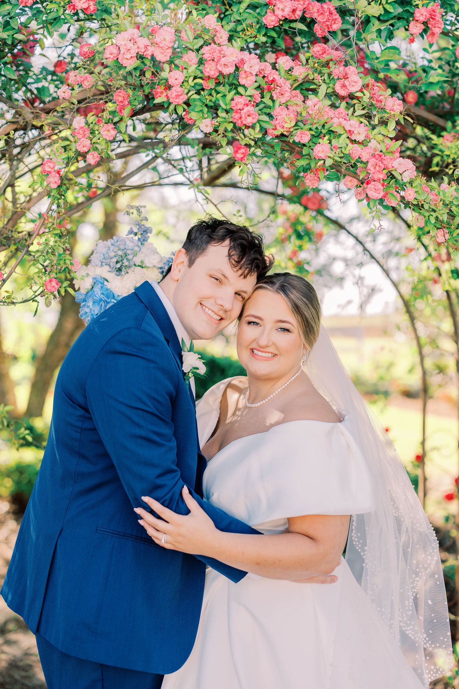 A romantic wedding photograph shows a dip kiss pose between newlyweds surrounded by greenery and tall grass at sunset.