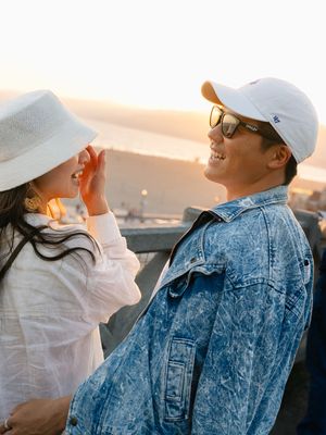 Smiling couple at sunset overlook, woman adjusting hat, man in denim jacket and white cap with sunglasses.