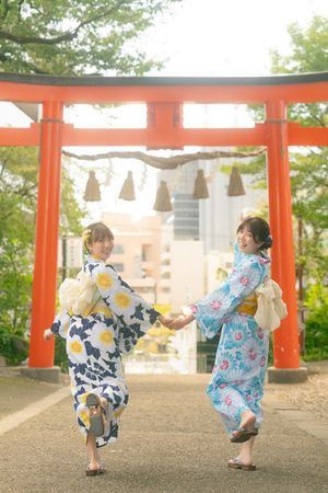 Two women in floral yukatas hold hands under a red torii gate at a Japanese shrine.