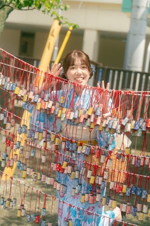 Young woman in blue floral yukata smiling behind colorful hanging Tanabata wish strips at Japanese festival.