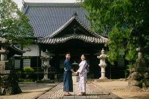Couple in traditional Japanese kimono standing before a historic temple with stone lanterns and lush trees.
