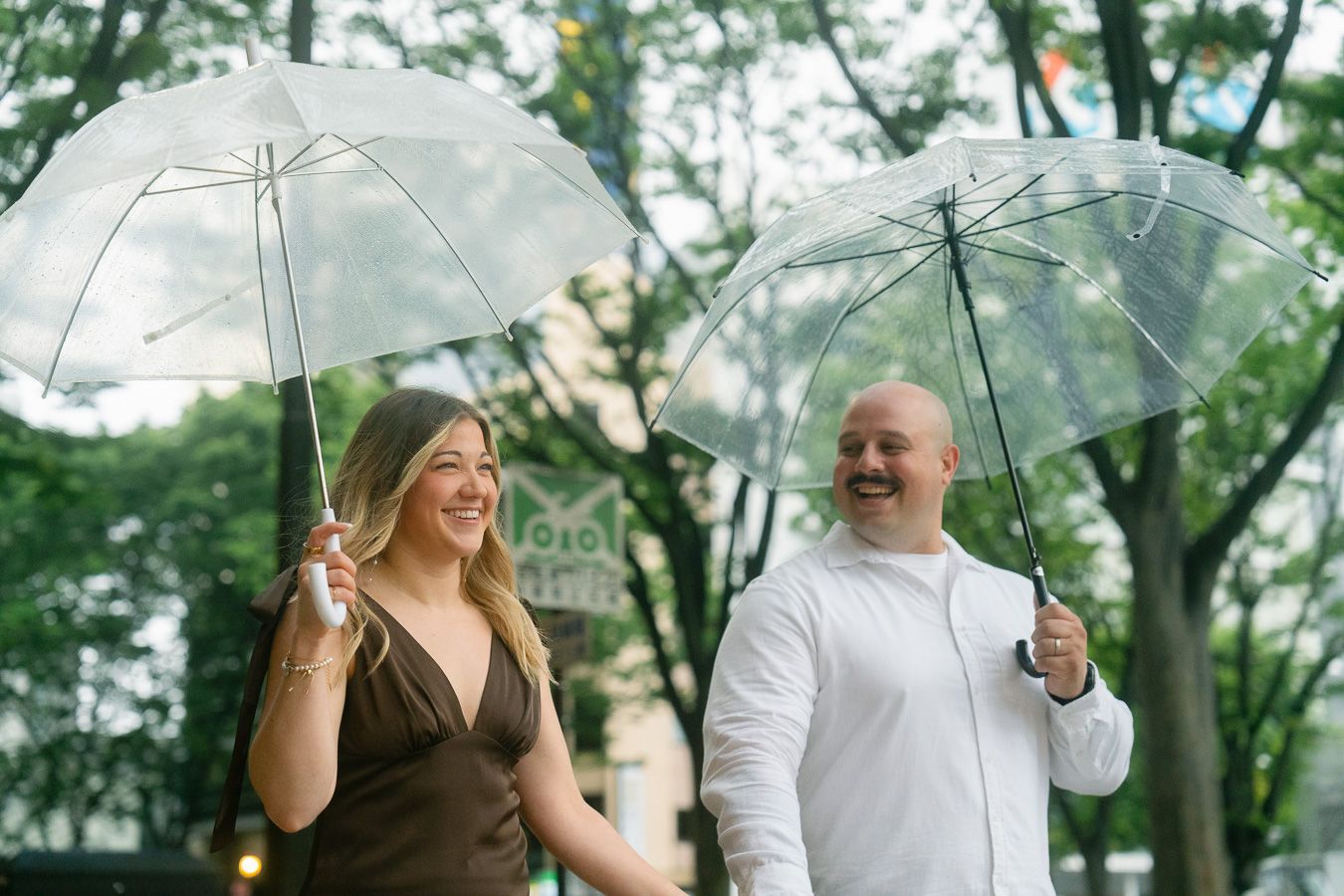 Couple smiling and walking under clear umbrellas in a lush green park on a rainy day.