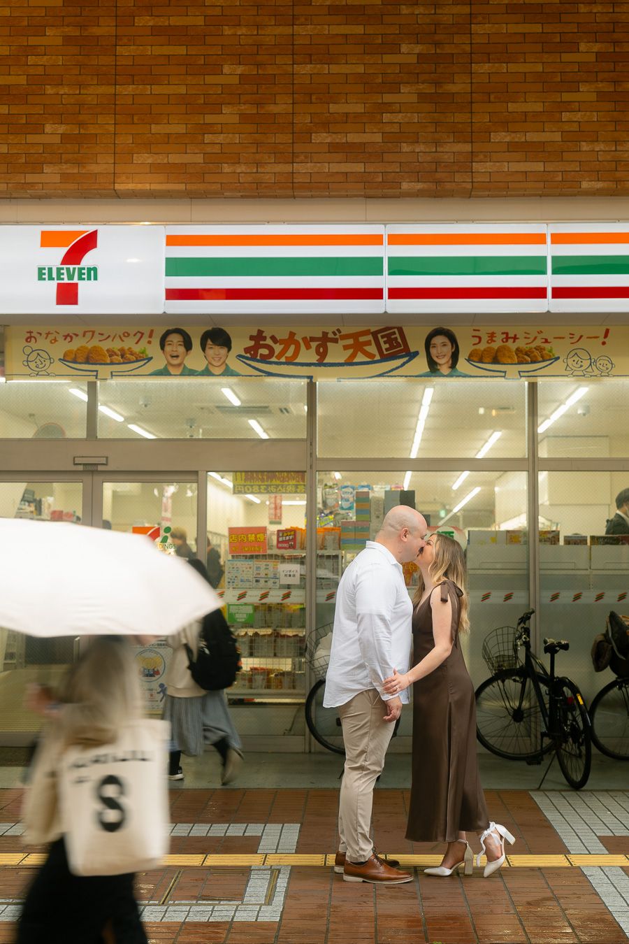 Couple kissing in front of a 7-Eleven convenience store in Japan, with passersby holding umbrellas nearby.