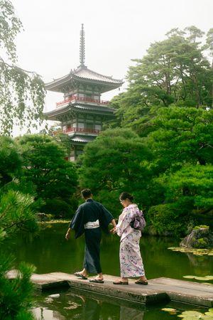 Two people in traditional Japanese kimonos walk on a wooden bridge by a serene pond with a pagoda and lush trees behind.