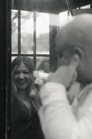 Black and white photo of a laughing woman holding a phone receiver inside a vintage phone booth with a man facing her.