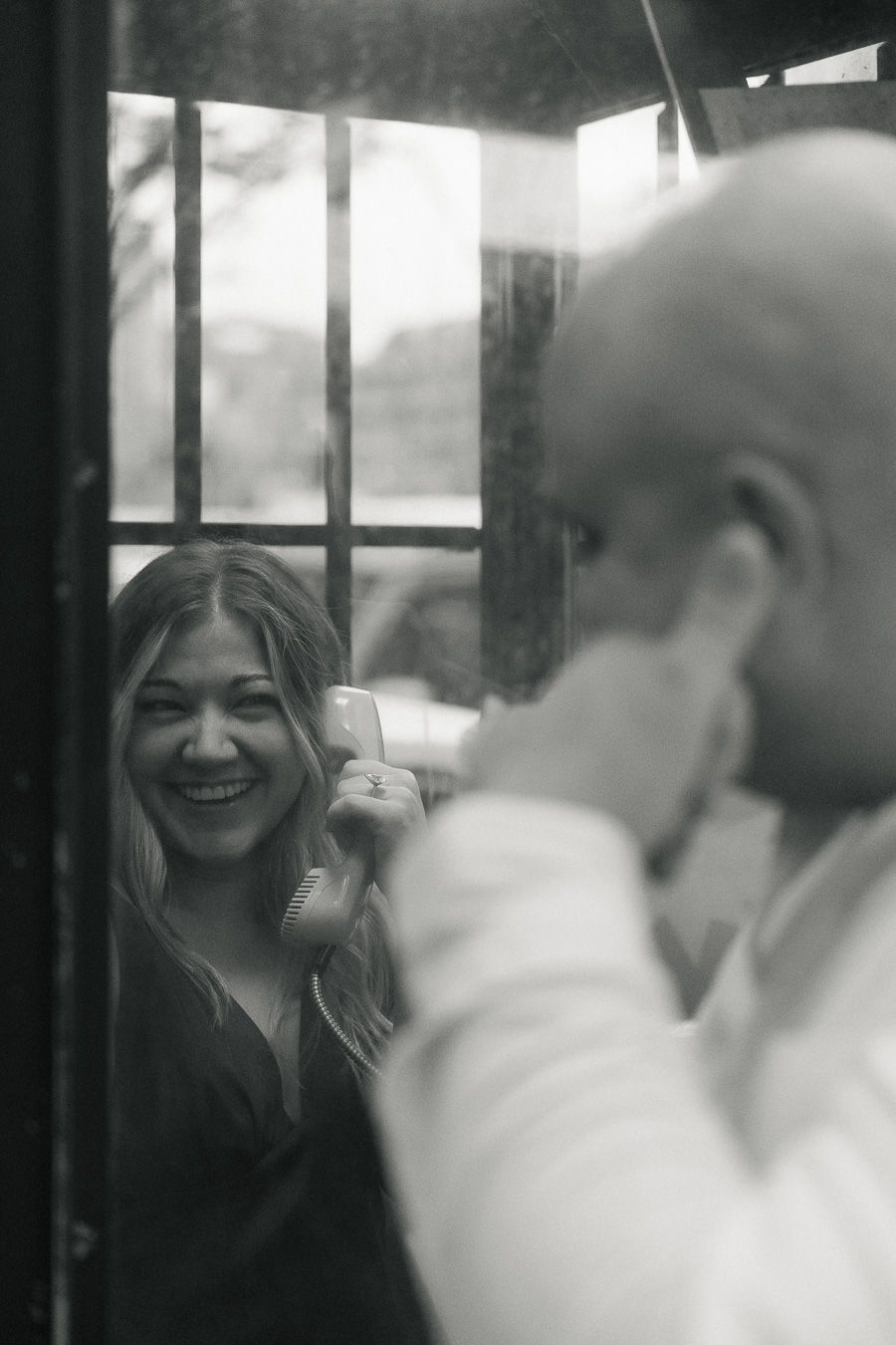 Black and white photo of a laughing woman holding a phone receiver inside a vintage phone booth with a man facing her.