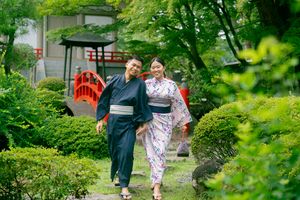 Couple wearing traditional Japanese kimono walking through a lush green garden with a red bridge backdrop.