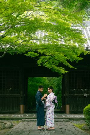 Couple in traditional Japanese kimono standing on stone path under lush green maple trees by wooden temple gate.