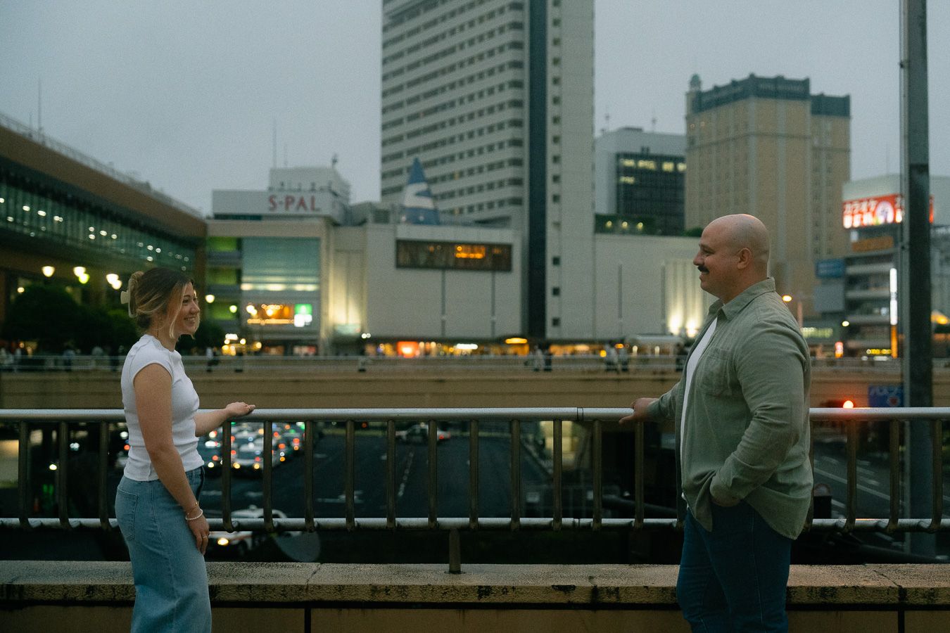 Two people stand apart on an urban rooftop terrace with city buildings and elevated train tracks in background.