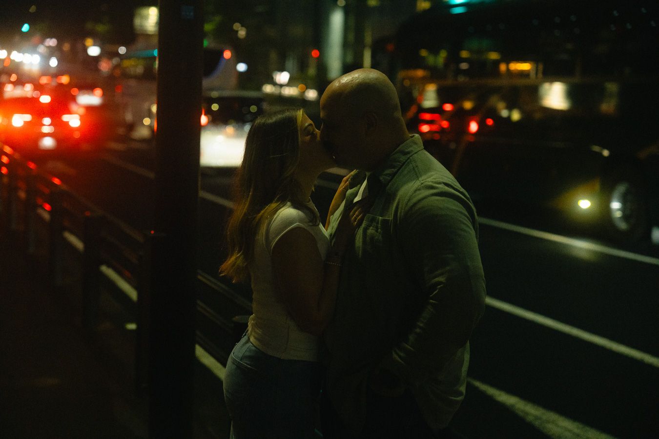 Couple sharing a romantic kiss on a city street at night with blurred car lights and traffic in the background.