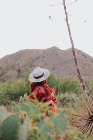 Fashion portrait in desert landscape featuring straw hat and red dress among cacti.