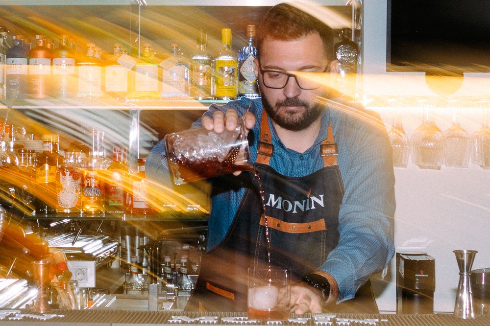 A bartender in denim overalls and gray sweatshirt pours a cocktail at a bar with illuminated bottles in the background.