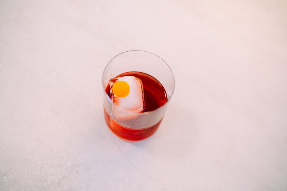 Red cocktail served in a rocks glass with a large spherical ice cube garnished with citrus peel.